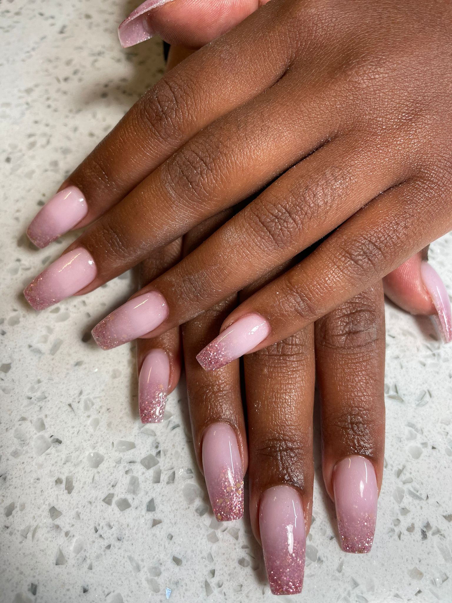 Roze glitter ombre nagels in coffin-vorm op de witte terrazzo balie van NAILS 72